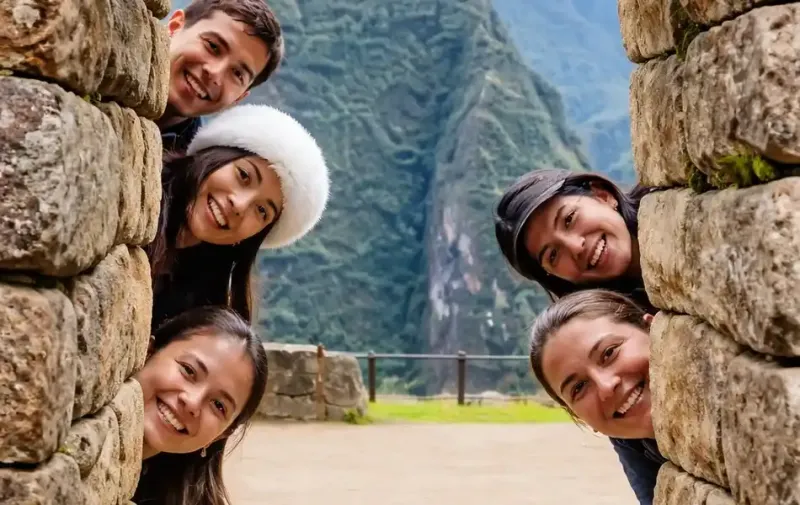 Grupo de amigos sonriendo detrás de un muro de piedra inca en la ciudadela de Machu Picchu | Inca Culture Travel